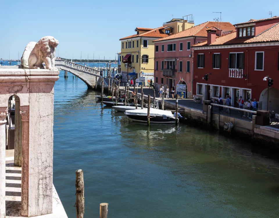 Scorcio di Canal Vena a Chioggia con il celebre Ponte di Vigo sullo sfondo, la statua del leone marciano in primo piano a sinistra e case storiche colorate lungo il canale.