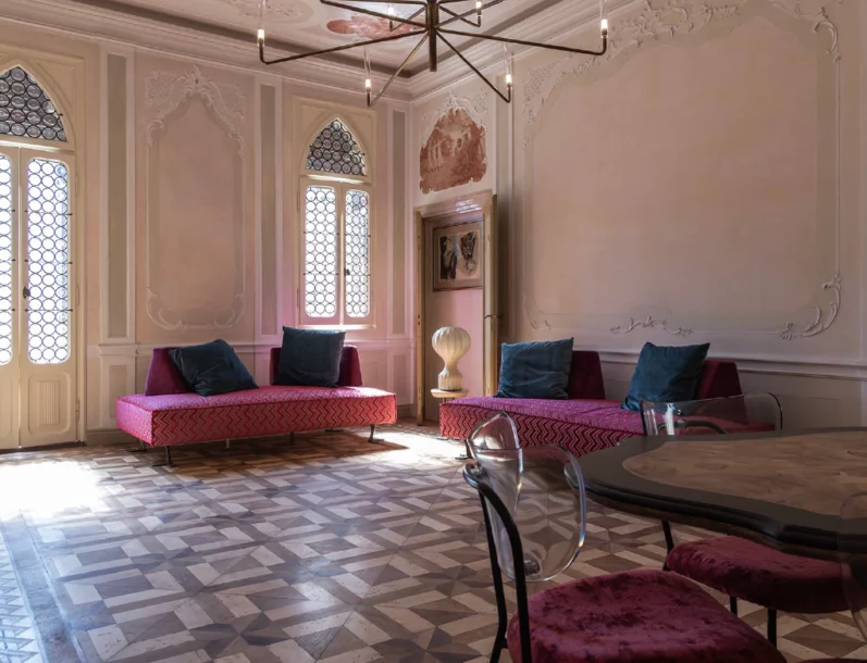 Ampio salone storico di Suite Attilia a Chioggia con pavimenti in legno intarsiato, due divani rossi geometrici e finestre veneziane ad arco.
