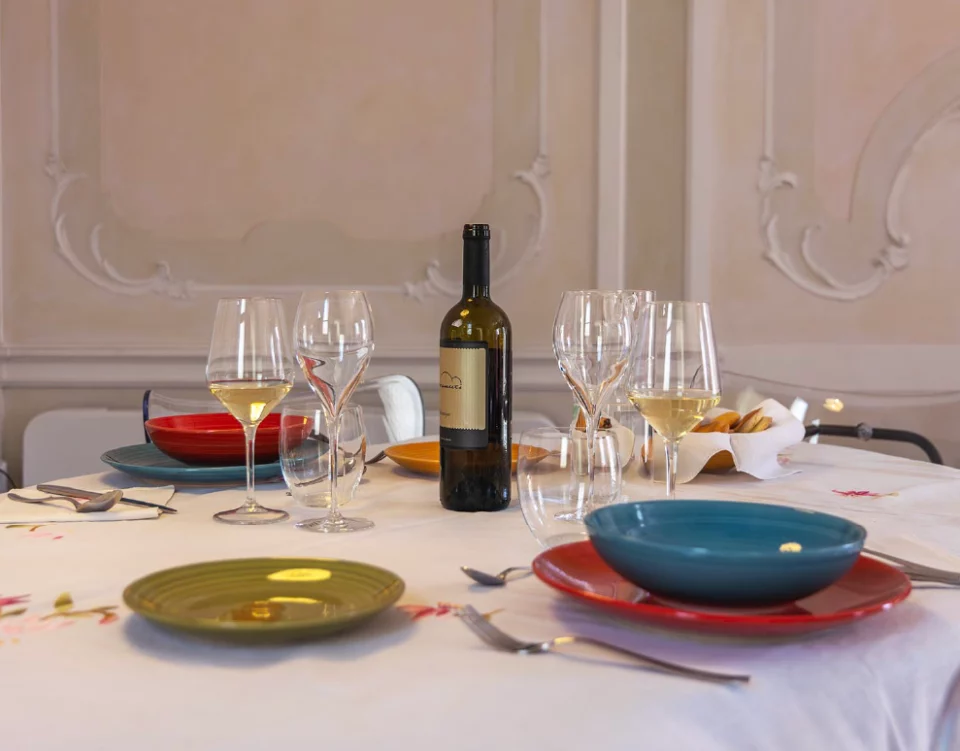 Tavola elegantemente apparecchiata con calici, piatti colorati e bottiglia di vino bianco nel salone con stucchi di Suite Attilia a Chioggia.